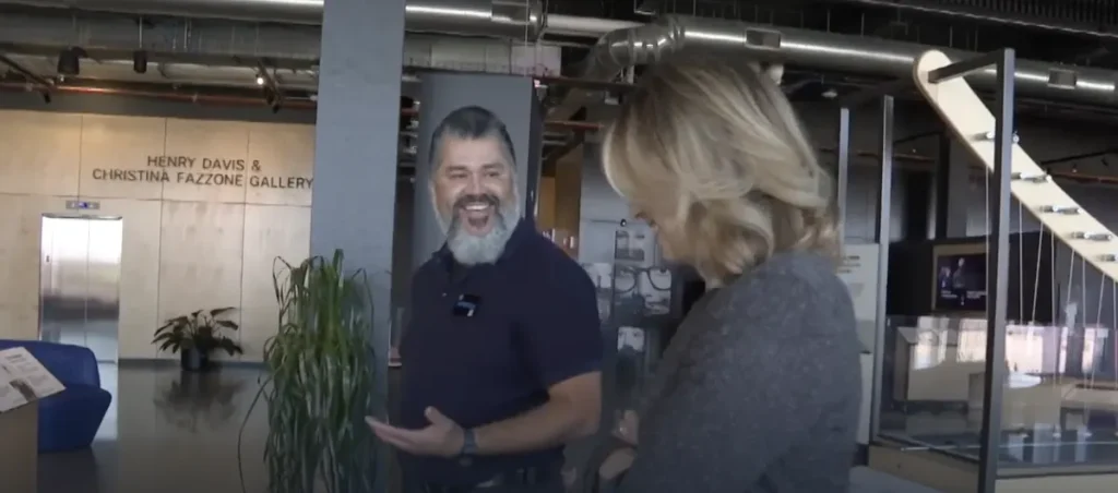 Anthony Galdamez, a Kiewit Luminarium staff member, smiles and gestures while speaking with KMTV reporter Mary Nelson, who laughs in response. They walking in the "Catch Waves" gallery, with a plant and exhibit elements visible in the background.